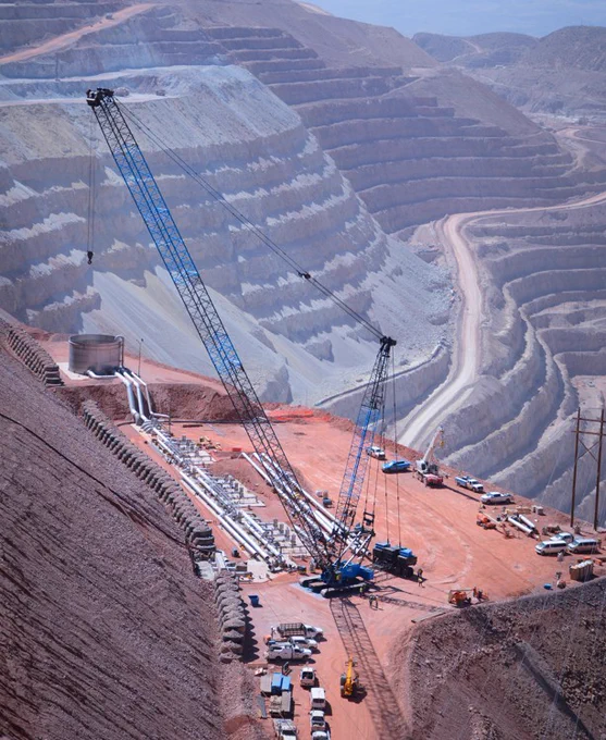 A large open-pit mine with terraced levels is shown. A large crane and various construction vehicles are on the reddish dirt surface, alongside pipes and equipment. The scene is expansive, with winding paths leading through the mine.