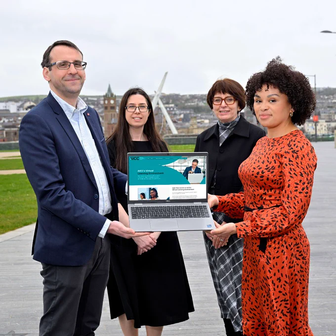 Four professionals standing outdoors by Derry~Londonderry waterfront, two holding a laptop displaying a website homepage.