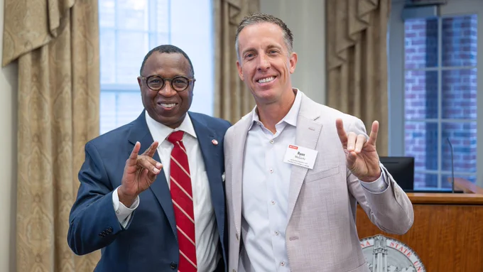 Chancellor Kevin Howell with Lenovo's Ryan McCurdy posing indoors, each making a 'wolfie' hand gesture while smiling for the camera.