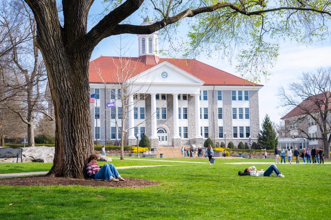 View of Wilson Hall in the back with students on the quad. A tree in the foreground has a student sitting and leaning against it.