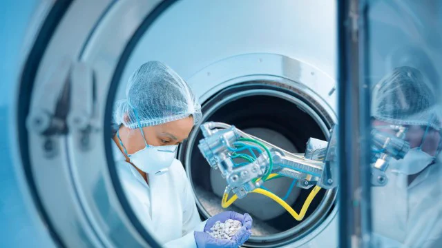 A pharmaceutical factory worker inspecting pills next to industrial manufacturing machinery. 