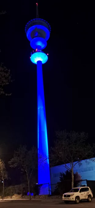 Descripción de la imagen: Foto nocturna de la alta torre de Telecom CLM, iluminada por completo de un color azul intenso y brillante. En primer plano a la derecha, se ve un vehículo todoterreno blanco de la empresa con el logo visible.