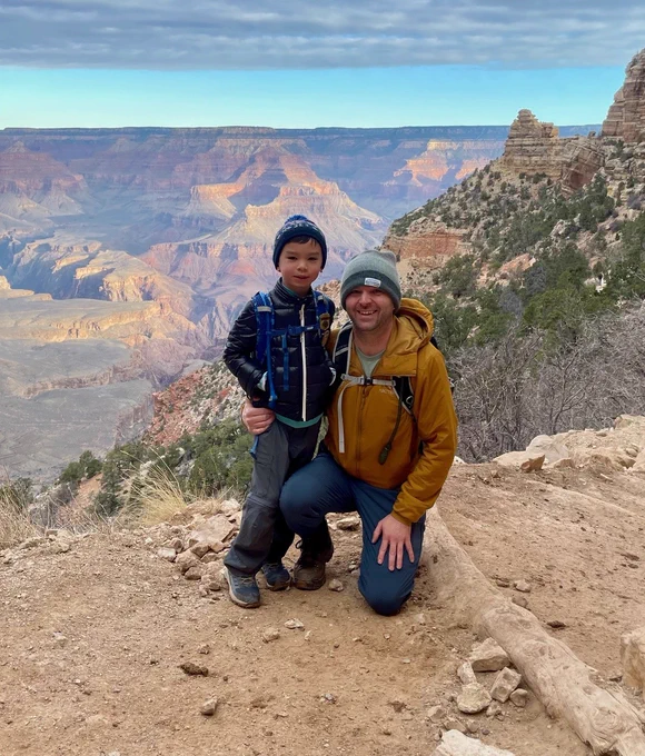 A man kneels with his arm around a child in front of the Grand Canyon. The man wears a yellow jacket and beanie, and the child wears a blue puffy jacket and hat. The Grand Canyon stretches behind them under a sky with clouds.