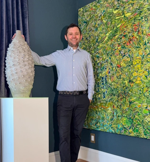 A smiling man in a striped shirt and dark pants poses beside a large vase covered in eye-like details. An abstract green and yellow painting hangs behind him, adding vibrant color to the dark blue wall.