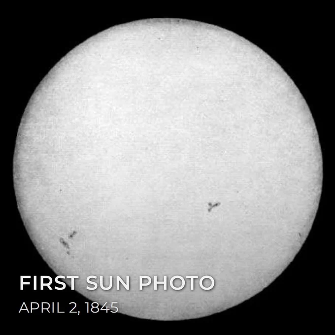 The first photograph of the Sun, a daguerreotype captured on April 2, 1845. The grainy black-and-white image shows a bright solar disk against a black background with dark sunspots visible on the lower half. Text overlay reads First Sun Photo, April 2, 1845.