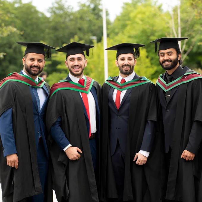 Four male graduates wearing black gowns and mortarboards stand together outdoors with greenery in the background.