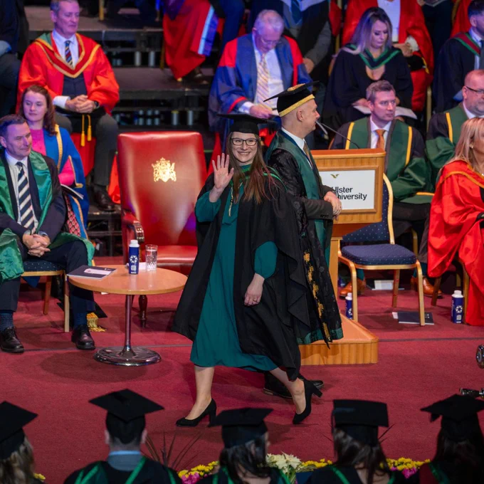 Graduate in green gown and black cap walking past a podium during a university ceremony with seated faculty in colourful academic robes.