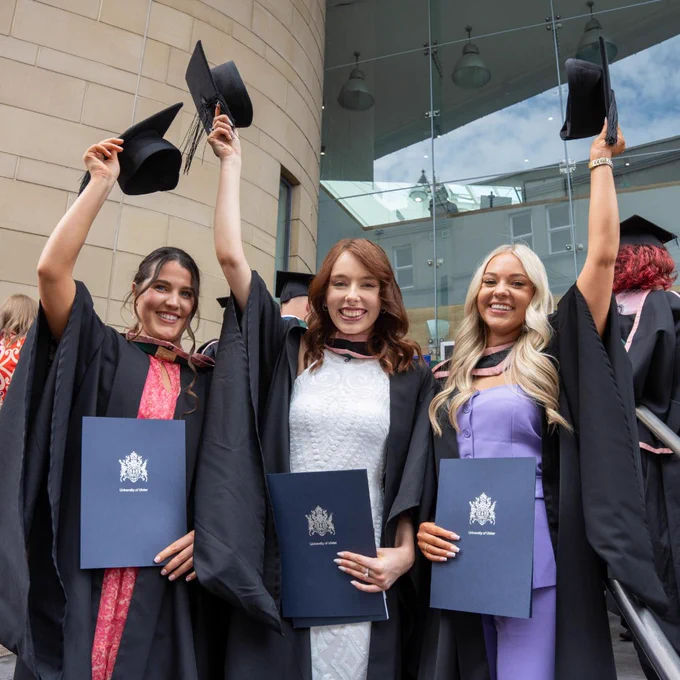 Three female graduates in black gowns and caps celebrate outside a modern building, holding diplomas and raising their caps in the air.