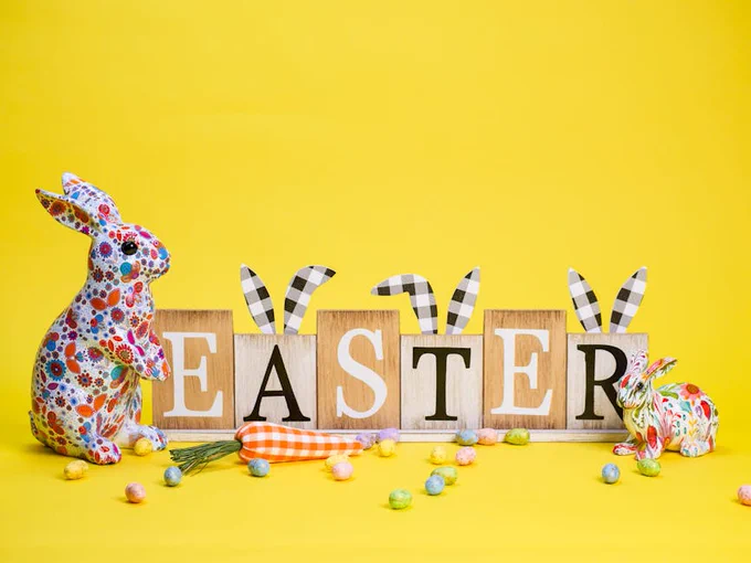 Colorful floral-patterned bunny figurines flank wooden blocks spelling EASTER against a bright yellow background with scattered pastel eggs and a fabric carrot.