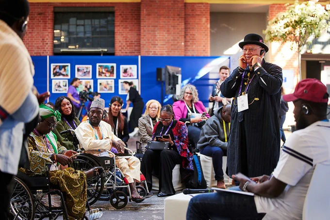 A diverse group of participants sits and listens during a session at GDS, while one person stands in the centre speaking and gesturing. Some participants are seated in wheelchairs, and others wear headsets.