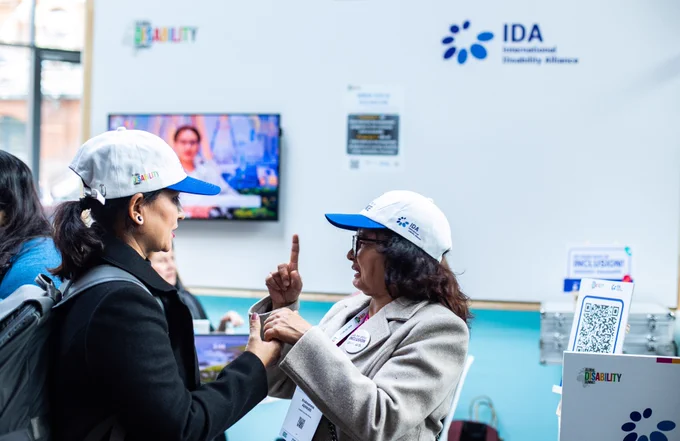 Two women wearing Global Disability Summit and IDA caps engaged in a conversation using sign language at an event booth, with informational displays and a QR code stand visible in the background.