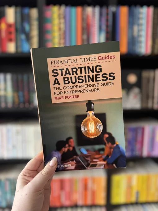 Hand holding the book Starting a Business: The Comprehensive Guide for Entrepreneurs by Mike Foster from the Financial Times Guides series, photographed in front of a colourful blurred bookshelf background.