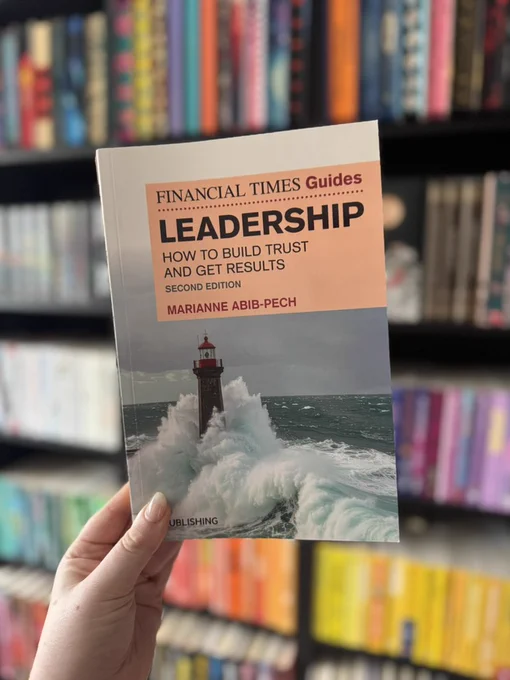 Hand holding the book Leadership: How to Build Trust and Get Results (Second Edition) by Marianne Abib Pech from the Financial Times Guides series, photographed in front of a blurred colourful bookshelf background.