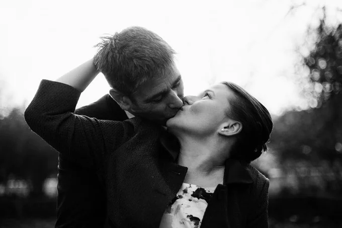 Schwarz/weiẞ Portrait einer jungen Frau, die von einem Mann geküsst wird, der hinter ihr steht. Mann sieht den Kopf der Frau im Profil. Sie steht rechts und dreht den Kopf nach Oben und zur Seite, um den Mann, der einen Kopf größer ist und sich zu ihr hinunterbeugt, auf dem Mund zu küssen. Dabei legt sie ihren rechten Arm um seinen Kopf. Im Hintergrund des im Freien aufgenommen Bildes sieht man unscharf Bäume.
