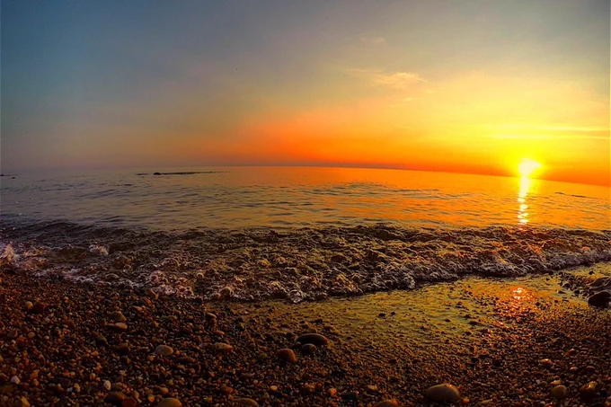 Sunset view over a lake from a pebble beach, with vibrant orange and yellow hues in the sky and sunlight reflecting on the water.