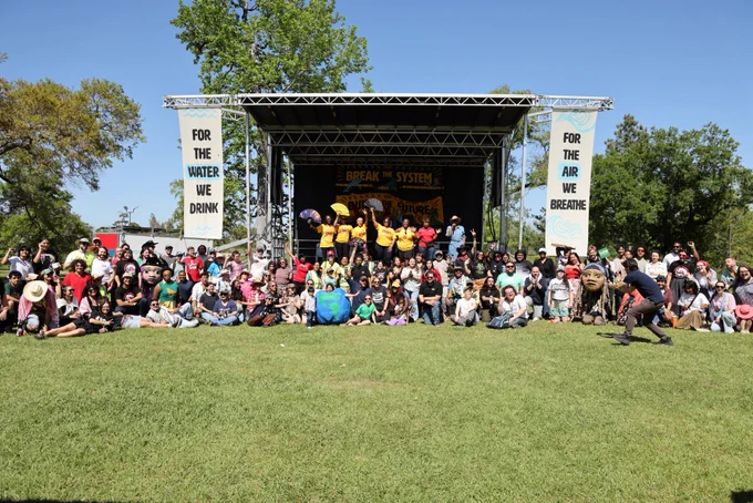 A crowd of community leaders before a stage hung with banners that say "For the water we drink" and "For the air we breathe." On stage, line dancers in yellow hold up fans. 