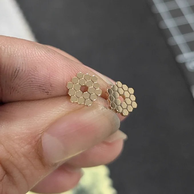 Two tiny 14K gold JWST mirror stud earrings held between fingertips, inspired after the James Webb Space Telescope primary mirror pattern
