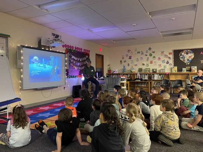A classroom setting with a large projection screen displaying educational content, surrounded by a group of attentive students sitting on the floor and listening to the presentation.