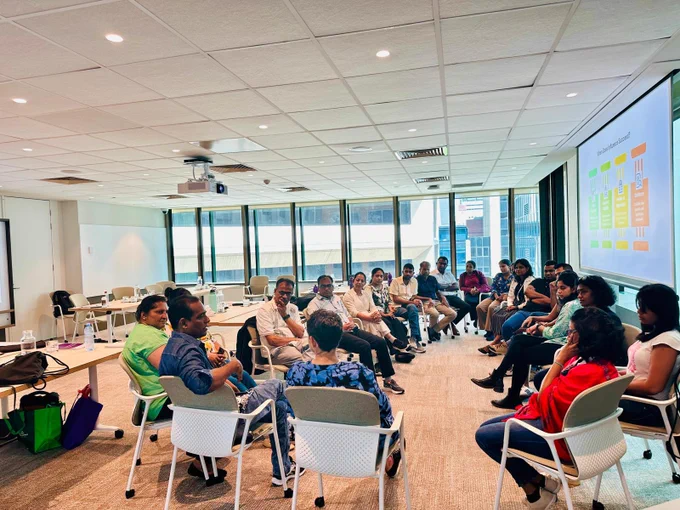 Participants seated in a circle during a collaborative workshop in a bright conference room with a presentation projected on the wall.