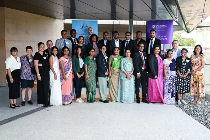 Group photo of in formal and traditional attire standing outside a modern building under a glass canopy with the Australia Awards banner and UQ International Development banner in the background. 