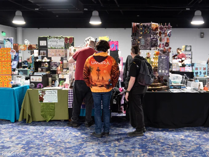 A photo of the 2025 Furry Migration Dealer's Den. There are several booths set up, with three attendees looking at one of them. 