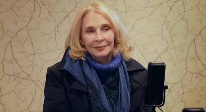 Gates McFadden wearing a black coat, with purple/blue scarf and jumper. Softly smiling.