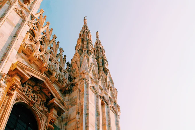 Upward view of the ornate Gothic façade of the Milan Cathedral (Duomo di Milano), showing detailed stone carvings, statues and pointed spires. The structure is illuminated by warm light against a clear, pale blue sky.
