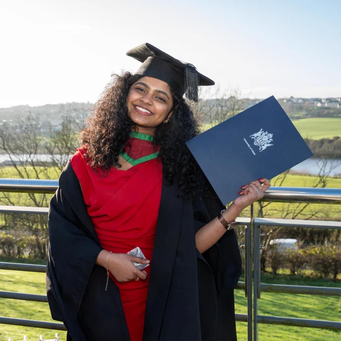 Graduate in cap and gown holding diploma outdoors with Coleraine campus river and fields in background.