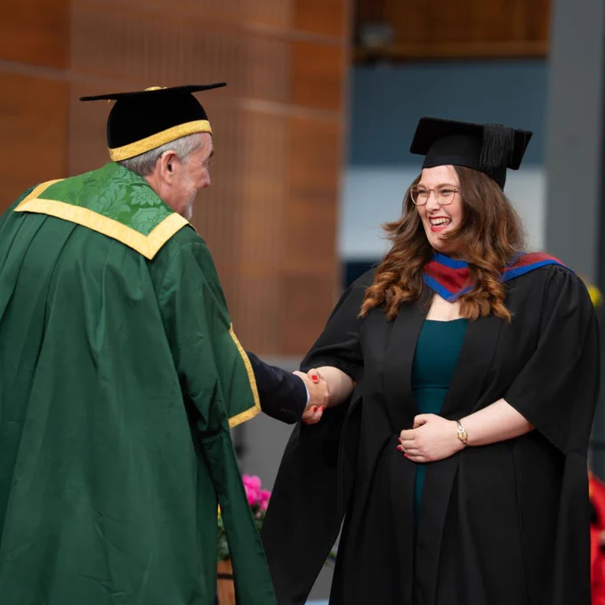 Graduate in black cap and gown shaking hands with Ulster University Deputy Vice Chancellor in green academic robe during ceremony.