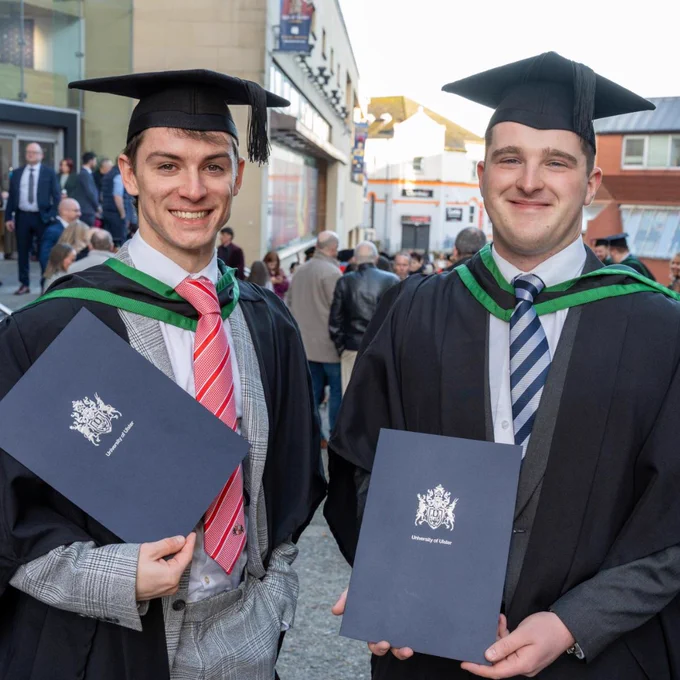 Two graduates in caps and gowns holding diploma folders outdoors with people and buildings in the background.