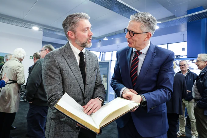 Two people stand in a busy room, looking at an open book held between them.