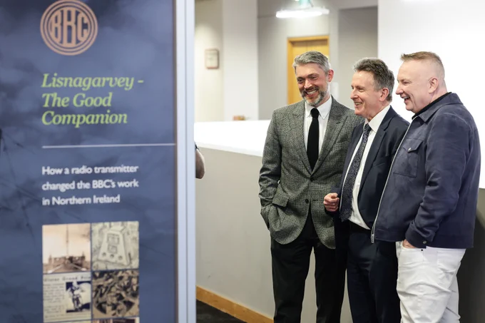 Three people stand together beside an exhibition panel titled “Lisnagarvey – The Good Companion.”