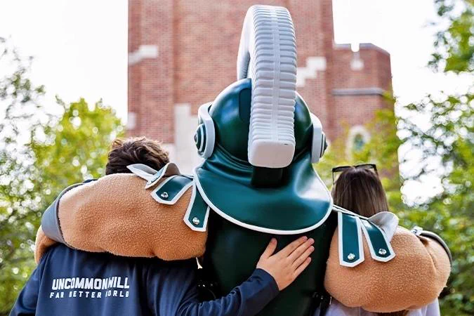 A Spartan mascot stands embraced by two people, one wearing a navy shirt with \"UNCOMMONHILL FAR BETTER WORLD\" text. The brick building and greenery in the background add depth to the image.