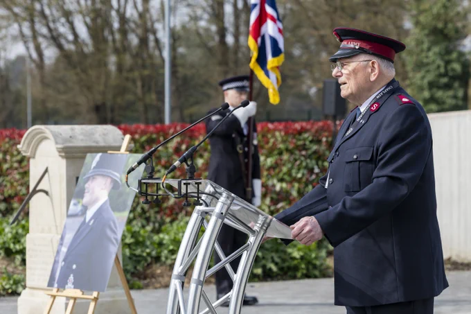 A uniformed speaker stands at a lectern addressing attendees, with an easel displaying a portrait of a uniformed officer beside them.