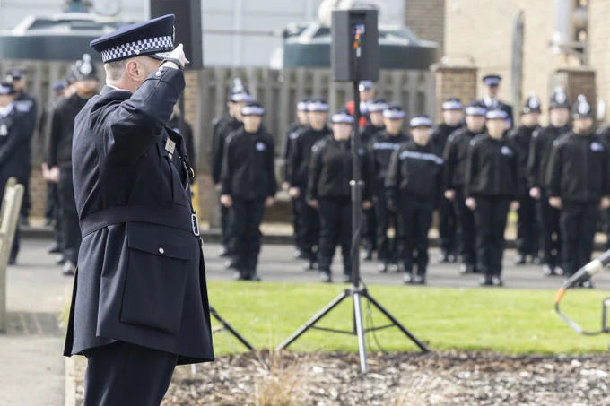 Chief Constable Tim Smith stands in the foreground saluting, while a formation of officers and staff stand lined up outdoors in the background.