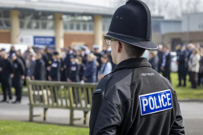 A uniformed officer is seen from behind watching a large gathering of officers and staff assembled outside a building.
