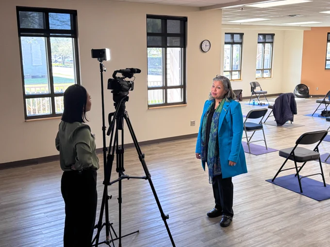 Karen Hua with CBS News stands behind a camera with Dr. Sallam in front of it answering interview questions. Behind Dr. Sallam is an open classroom with folding chairs arranged on top of yoga mats. 