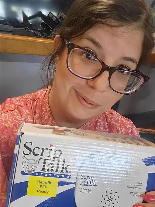 Jenna is holding a ScripTalk Reader and ready to present. She's a brunette with her hair pulled back and wearing dark glasses and a pink blouse. 