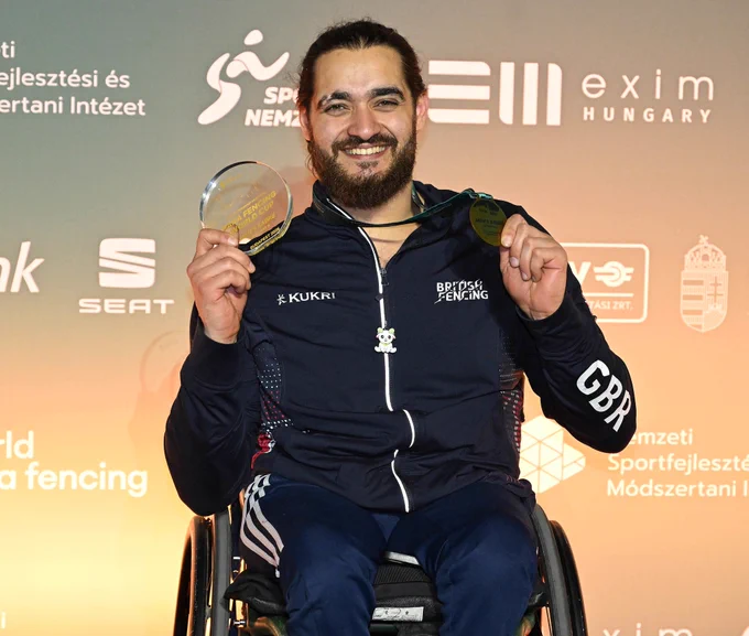 A GBR para fencer smiles to the camera, holding up a trophy and gold medal.