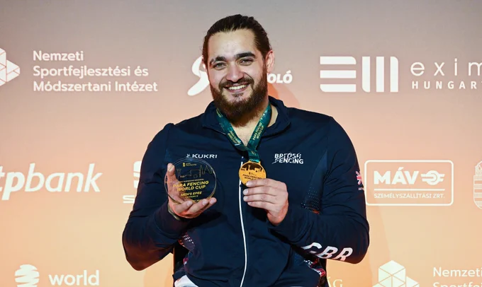 A GBR para fencer smiles to the camera, holding up a trophy and gold medal.
