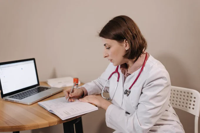 A doctor in a white coat with a stethoscope around her neck is sitting at a wooden table. She is writing notes in a notebook, with an open laptop beside her.