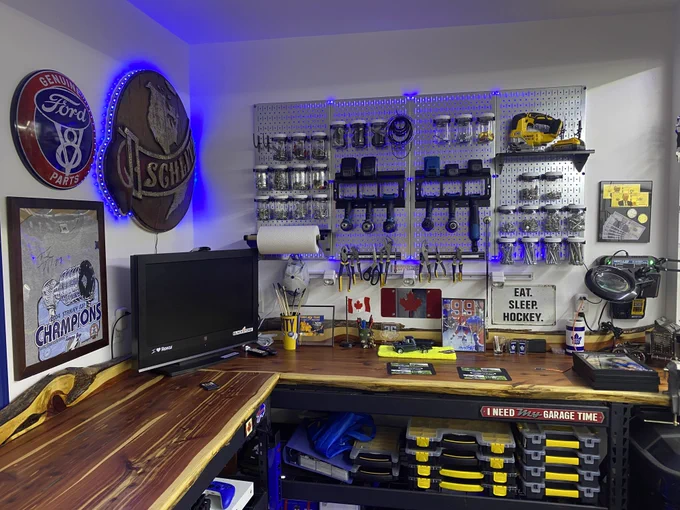 Wall Control metal pegboard mounted above a garage workbench, organized with jars of hardware, hand tools, and power tools, illuminated by blue LED lighting, with a wooden work surface, monitor, storage bins, and automotive-themed wall decor. Photo by customer John.