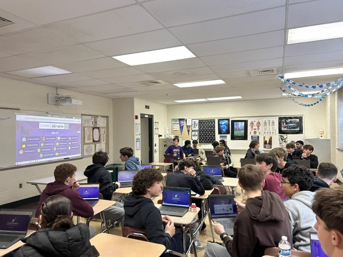High school students seated in a classroom using laptops while a quiz game is projected on the whiteboard at the front.