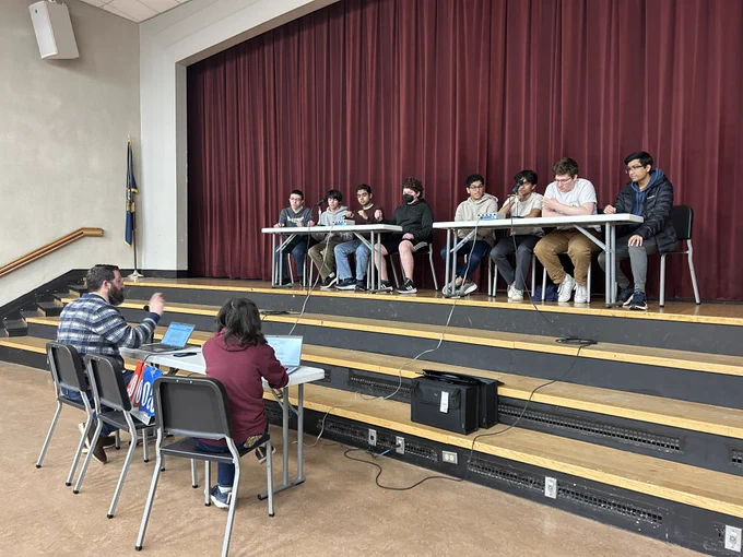 A group of eight young people seated at a long table on a stage, facing two individuals with laptops seated below them in a discussion setup.