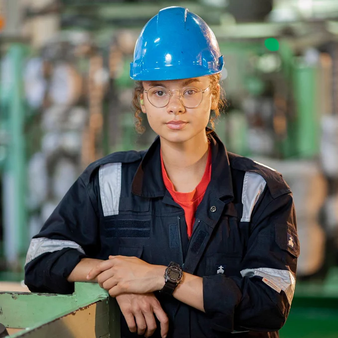A young woman in a blue hard hat and navy work coveralls with reflective strips stands confidently in an industrial setting. She is wearing glasses and a digital watch, leaning against green machinery with a blurred factory background.