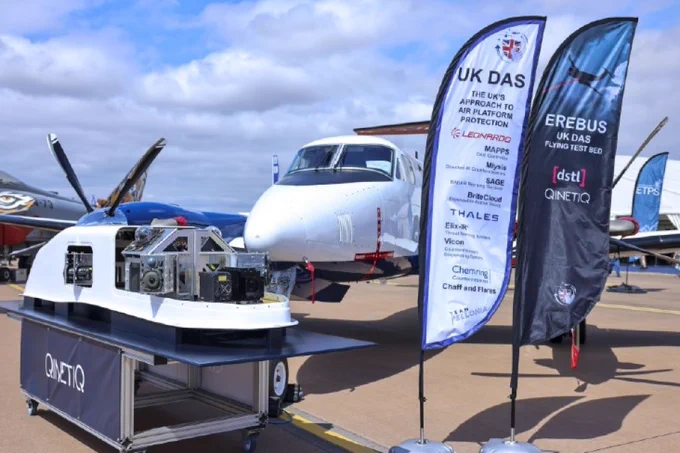 Small white aircraft parked on tarmac with two tall flags displaying UK DAS and EREBUS/Dstl/QinetiQ branding beside a technical equipment setup on a table.