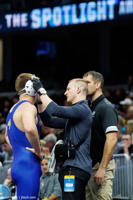 An athletic trainer patches up a wrestler in a blue singlet's nose while a coach in a black polo looks on.