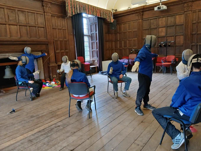 Several people in fencing gear are doing seated fencing in a room. Some are refereeing.