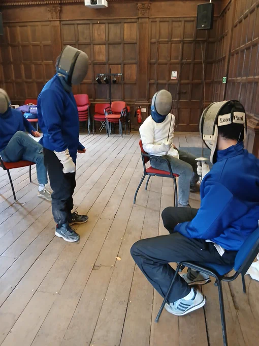 Several people in fencing gear are doing seated fencing in a room. Some are refereeing.