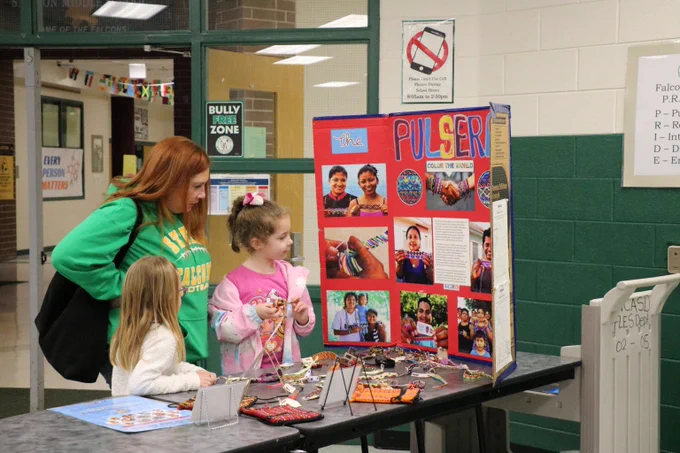 Three children examine a colorful project display titled 'PULSER' set up on a table in a school hallway.